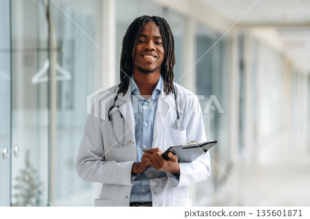 Joyful, positive facial expression. Young male black doctor is in the corridor Joyful, positive facial expression. Young male black doctor is in the corridor 135601871