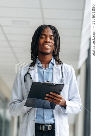 Stethoscope on neck, notepad in hands. Young male black doctor is in the corridor 135601880