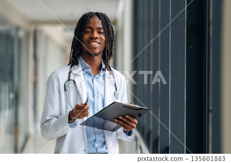 Stethoscope on neck, notepad in hands. Young male black doctor is in the corridor Stethoscope on neck, notepad in hands. Young male black doctor is in the corridor 135601883