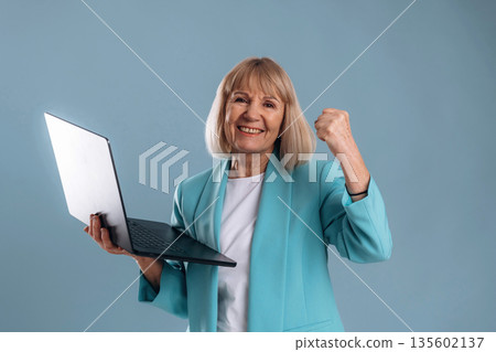 Celebrating success, holding laptop. Senior woman is against background Celebrating success, holding laptop. Senior woman is against background 135602137
