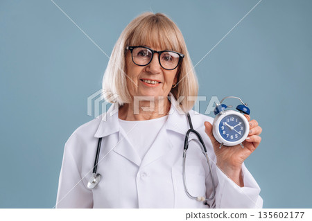 Clock in hand, smiling. Senior woman doctor is against background Clock in hand, smiling. Senior woman doctor is against background 135602177