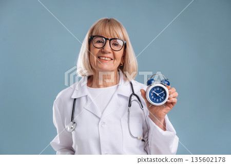 Clock in hand, smiling. Senior woman doctor is against background Clock in hand, smiling. Senior woman doctor is against background 135602178