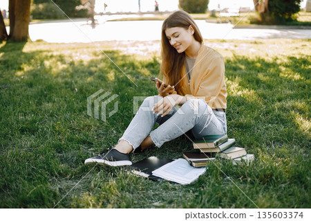 Young girl using a smartphone while sitting on a grass. Woman have a books around her. Girl wearing a beige shirt and jeans. Young girl using a smartphone while sitting on a grass. Woman have a books around her. Girl wearing a beige shirt and jeans. 135603374
