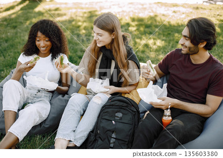 Friends hanging in the green park, relaxing. Boy and girls holding a bottles of juice. African girl wearing white clothes, boy wearing brown t-shirt and black jeans. Friends hanging in the green park, relaxing. Boy and girls holding a bottles of juice. African girl wearing white clothes, boy wearing brown t-shirt and black jeans. 135603380