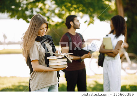 Focus on a caucasian girl who standing sepately. Group of international students standing together in park at university. Blurred african girl and indian boy talking outdoors. 135603399