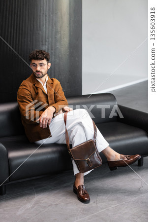 Elegant man in a brown corduroy blazer and white trousers sitting on a black leather sofa with a premium dark brown leather messenger bag in a minimalist studio. 135603918