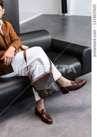 Elegant man in a brown corduroy blazer and white trousers sitting on a black leather sofa with a premium dark brown leather messenger bag in a minimalist studio. Elegant man in a brown corduroy blazer and white trousers sitting on a black leather sofa with a premium dark brown leather messenger bag in a minimalist studio. 135603920