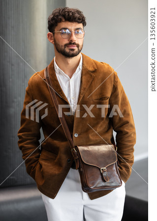 Portrait of a stylish man in a brown corduroy blazer and white shirt holding a black leather messenger bag in a minimalist studio. 135603921