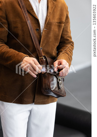 Close-up of a man in a brown corduroy jacket holding a brown leather messenger bag with a buckle closure in a minimalist studio. 135603922