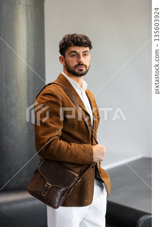 Portrait of a stylish man in a brown corduroy blazer and white shirt holding a cognac leather messenger bag in a minimalist studio. Portrait of a stylish man in a brown corduroy blazer and white shirt holding a cognac leather messenger bag in a minimalist studio. 135603924