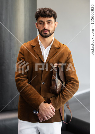Portrait of a stylish man in a brown corduroy blazer and white shirt holding a cognac leather messenger bag in a minimalist studio. Portrait of a stylish man in a brown corduroy blazer and white shirt holding a cognac leather messenger bag in a minimalist studio. 135603926