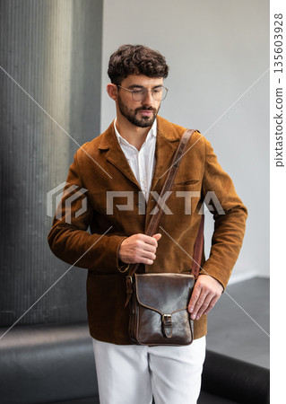 Portrait of a stylish man in a brown corduroy blazer and white shirt holding a cognac leather messenger bag in a minimalist studio. Portrait of a stylish man in a brown corduroy blazer and white shirt holding a cognac leather messenger bag in a minimalist studio. 135603928