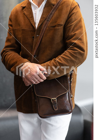 Close-up of a man in a brown corduroy jacket holding a brown leather messenger bag with a buckle closure in a minimalist studio. 135603932