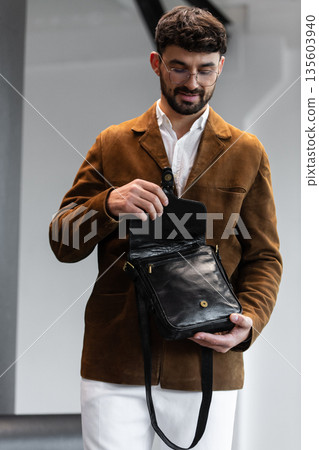 Portrait of a stylish man in a brown corduroy blazer and white shirt holding a black leather messenger bag in a minimalist studio. 135603940