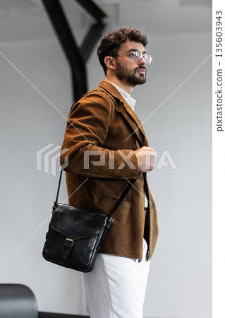 Portrait of a stylish man in a brown corduroy blazer and white shirt holding a black leather messenger bag in a minimalist studio. Portrait of a stylish man in a brown corduroy blazer and white shirt holding a black leather messenger bag in a minimalist studio. 135603943