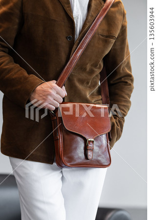 Close-up of a premium cognac leather messenger bag with a metal buckle held by a man in a brown corduroy blazer in a minimalist studio Close-up of a premium cognac leather messenger bag with a metal buckle held by a man in a brown corduroy blazer in a minimalist studio 135603944