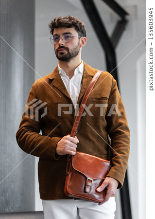 Portrait of a stylish man in a brown corduroy blazer and white shirt holding a cognac leather messenger bag in a minimalist studio. Portrait of a stylish man in a brown corduroy blazer and white shirt holding a cognac leather messenger bag in a minimalist studio. 135603945