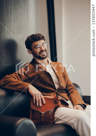 Sophisticated man in a brown corduroy blazer and glasses sitting on a black leather sofa holding a cognac leather messenger bag in a minimalist studio. Sophisticated man in a brown corduroy blazer and glasses sitting on a black leather sofa holding a cognac leather messenger bag in a minimalist studio. 135603947