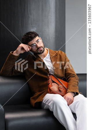 Sophisticated man in a brown corduroy blazer and glasses sitting on a black leather sofa holding a cognac leather messenger bag in a minimalist studio. Sophisticated man in a brown corduroy blazer and glasses sitting on a black leather sofa holding a cognac leather messenger bag in a minimalist studio. 135603967