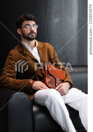 Sophisticated man in a brown corduroy blazer and glasses sitting on a black leather sofa holding a cognac leather messenger bag in a minimalist studio. Sophisticated man in a brown corduroy blazer and glasses sitting on a black leather sofa holding a cognac leather messenger bag in a minimalist studio. 135603974