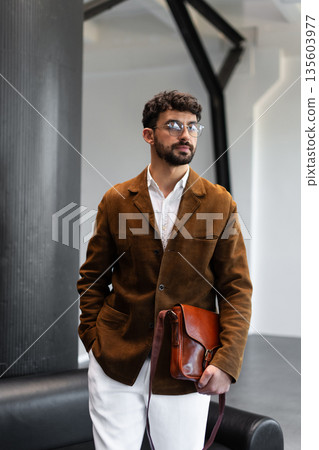 Portrait of a stylish man in a brown corduroy blazer and white shirt holding a cognac leather messenger bag in a minimalist studio. Portrait of a stylish man in a brown corduroy blazer and white shirt holding a cognac leather messenger bag in a minimalist studio. 135603977