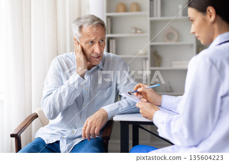 Senior male patient reporting ear pain to female doctor during medical visit Senior male patient reporting ear pain to female doctor during medical visit 135604223
