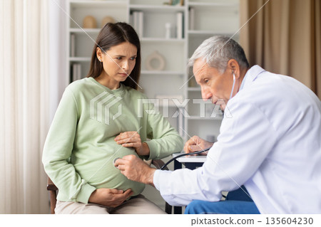 Frustrated senior male doctor examining pregnant woman with stethoscope during prenatal visit Frustrated senior male doctor examining pregnant woman with stethoscope during prenatal visit 135604230