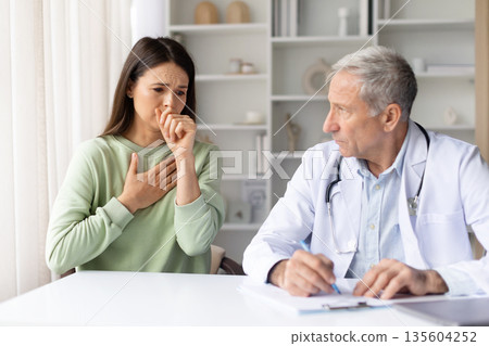 Female patient coughing during medical examination at clinic with senior male doctor Female patient coughing during medical examination at clinic with senior male doctor 135604252