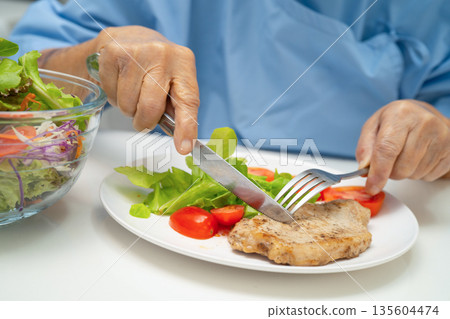 Asian elderly woman patient eating pork chop stake and vegetable salad for healthy food in hospital. 135604474
