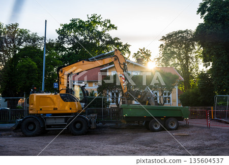 Road construction work with yellow excavator loading dump trailer at residential street during sunset 135604537