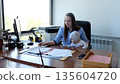 Businesswoman managing paperwork and childcare, holding her baby on her lap while working at her desk in a bright office 135604720