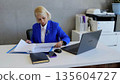 Focused businesswoman in blue suit reviewing documents at modern office desk with laptop and smartphone, showing professionalism and dedication 135604727