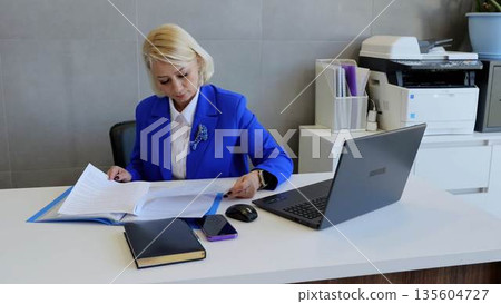 Focused businesswoman in blue suit reviewing documents at modern office desk with laptop and smartphone, showing professionalism and dedication Focused businesswoman in blue suit reviewing documents at modern office desk with laptop and smartphone, showing professionalism and dedication 135604727