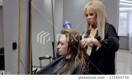 Hairdresser carefully drying and styling a client's hair in a cozy salon, creating a trendy look with expertise and precision Hairdresser carefully drying and styling a client's hair in a cozy salon, creating a trendy look with expertise and precision 135604730