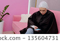 Woman sitting on a pink couch in a hair salon, wearing a disposable cap and browsing on her smartphone while waiting for her hair coloring treatment 135604731