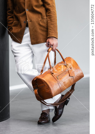 Close-up of a man holding a premium brown leather duffel bag wearing a suede blazer and white trousers 135604772