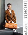 Stylish man in brown suede jacket and glasses leaning against a pillar while holding a leather weekend bag 135604854