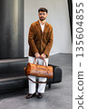 Handsome stylish man in brown suede blazer and white trousers holding premium leather duffel bag in modern interior 135604855