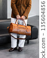 Close-up of a man holding a premium brown leather duffel bag wearing a suede blazer and white trousers 135604856
