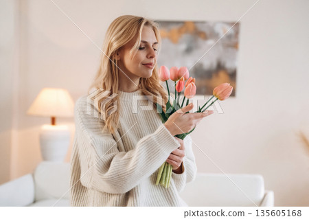 Admiring the gift, standing. Beautiful young woman is at home with flowers Admiring the gift, standing. Beautiful young woman is at home with flowers 135605168
