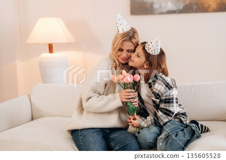 In birthday hats, celebrating. Mother and daughter are together in domestic room In birthday hats, celebrating. Mother and daughter are together in domestic room 135605528