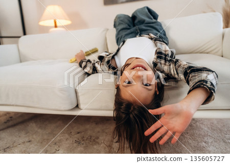 Upside down, lying down on the sofa. Cute little girl is in the domestic room alone Upside down, lying down on the sofa. Cute little girl is in the domestic room alone 135605727