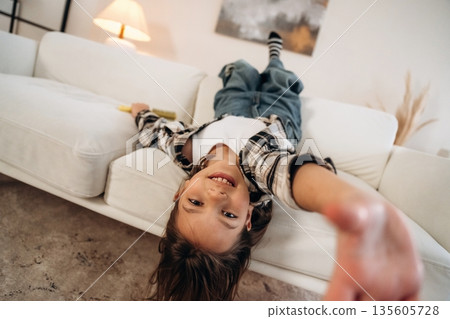 Upside down, lying down on the sofa. Cute little girl is in the domestic room alone Upside down, lying down on the sofa. Cute little girl is in the domestic room alone 135605728