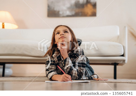 Drawing on the paper, lying down on floor. Cute little girl is in the domestic room alone Drawing on the paper, lying down on floor. Cute little girl is in the domestic room alone 135605741
