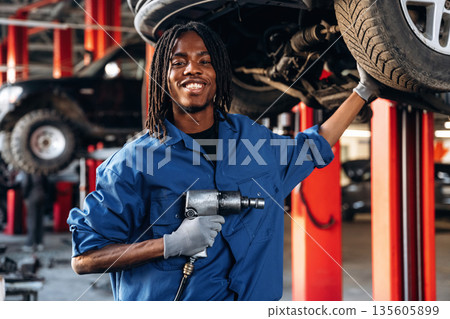 Vehicle is high up, about to change the tire. Male mechanic is with car in garage 135605899