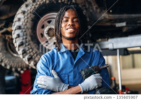 In blue uniform and with automatic screwdriver. Male mechanic is with car in garage In blue uniform and with automatic screwdriver. Male mechanic is with car in garage 135605907