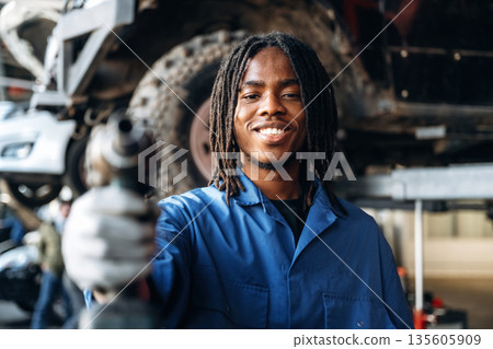 In blue uniform and with automatic screwdriver. Male mechanic is with car in garage In blue uniform and with automatic screwdriver. Male mechanic is with car in garage 135605909