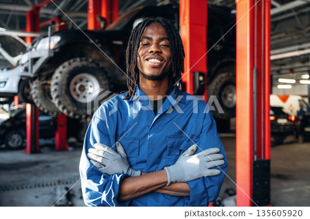 Dreadlocks hairstyle, in blue uniform. Male mechanic is with car in garage Dreadlocks hairstyle, in blue uniform. Male mechanic is with car in garage 135605920