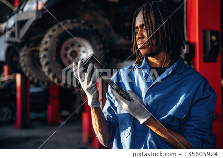 Automobile part in hands. Male mechanic is with car in garage Automobile part in hands. Male mechanic is with car in garage 135605924