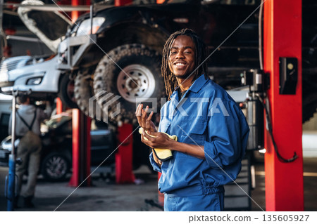 Dreadlocks hairstyle, in blue uniform. Male mechanic is with car in garage Dreadlocks hairstyle, in blue uniform. Male mechanic is with car in garage 135605927